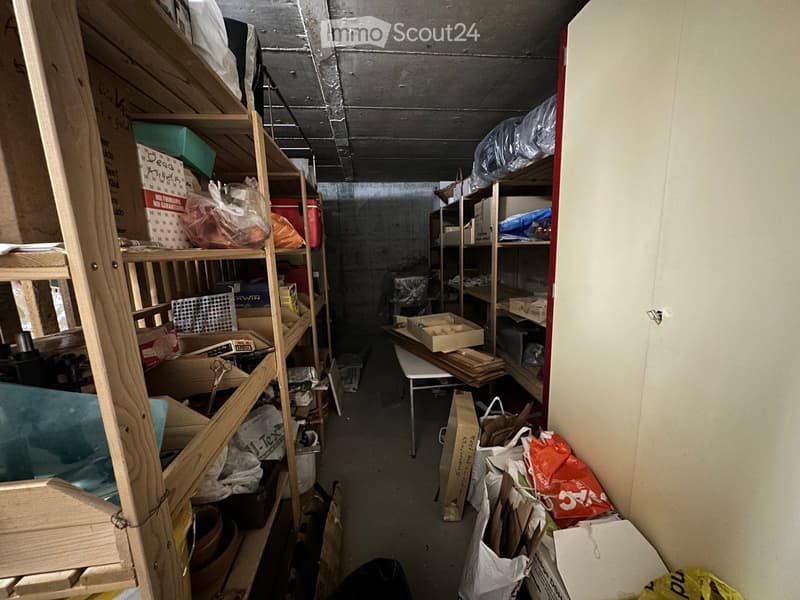 A cluttered storage area with wooden shelves filled with various items, including cardboard boxes, plastic bags, and wooden planks. A small table is positioned in the center, and the space has a concrete ceiling and walls.