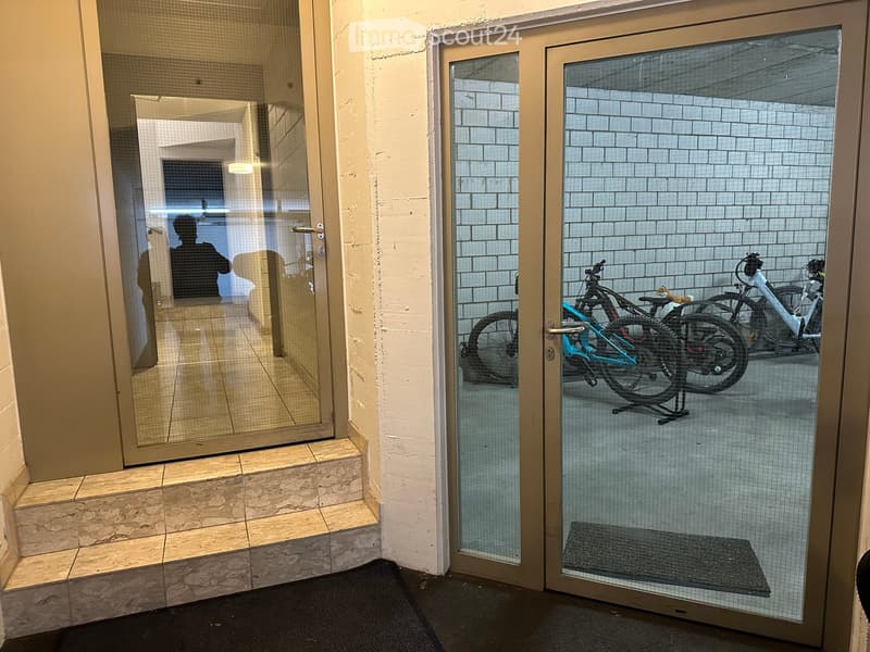 Hallway with glass doors leading to a bicycle storage area, featuring tiled floors and white brick walls.