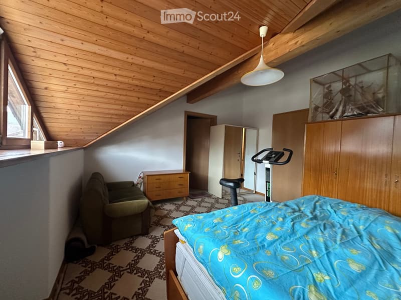 Cozy attic bedroom with wooden ceiling beams, a bed with blue bedding, a small sofa, an exercise bike, and built-in wooden wardrobes.