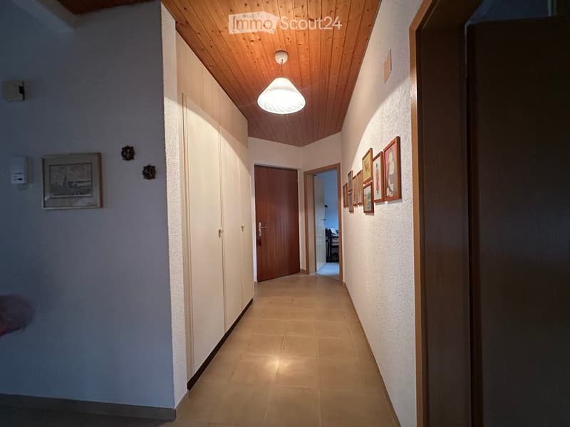 A narrow hallway with beige tiled flooring, white walls, and a wooden ceiling. Features include built-in white closet doors, framed artwork, and a hanging light fixture. A wooden door leads to another room.