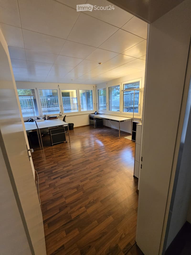 Bright office space with wooden flooring, large windows, and modern furniture including desks, chairs, and cabinets.