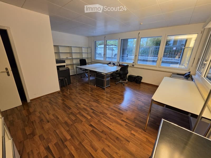 Office space with wooden flooring, large windows, white walls, and modern furniture including a desk, chairs, shelves, and a printer.