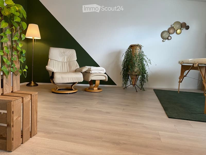 A modern, minimalistic room with light wooden flooring, a cream-colored armchair, a matching footstool, a floor lamp, a small table with a plant, and a decorative wall art piece.
