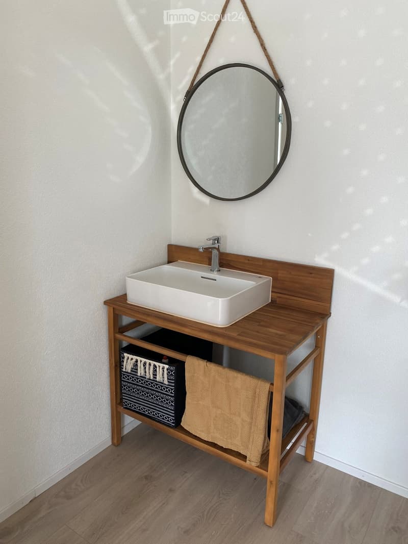 Bathroom with a wooden vanity, white sink, chrome faucet, round mirror, and a towel on the shelf below.