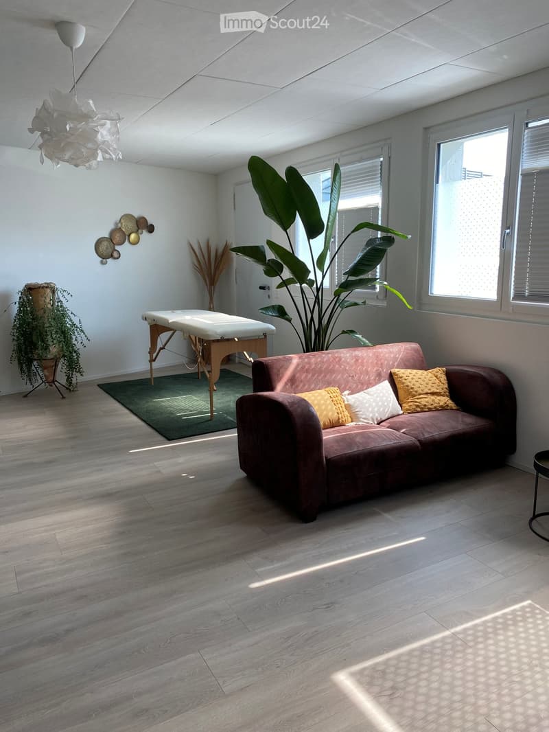 Modern living room with wooden flooring, large windows, a brown sofa with yellow pillows, a massage table, and decorative plants.