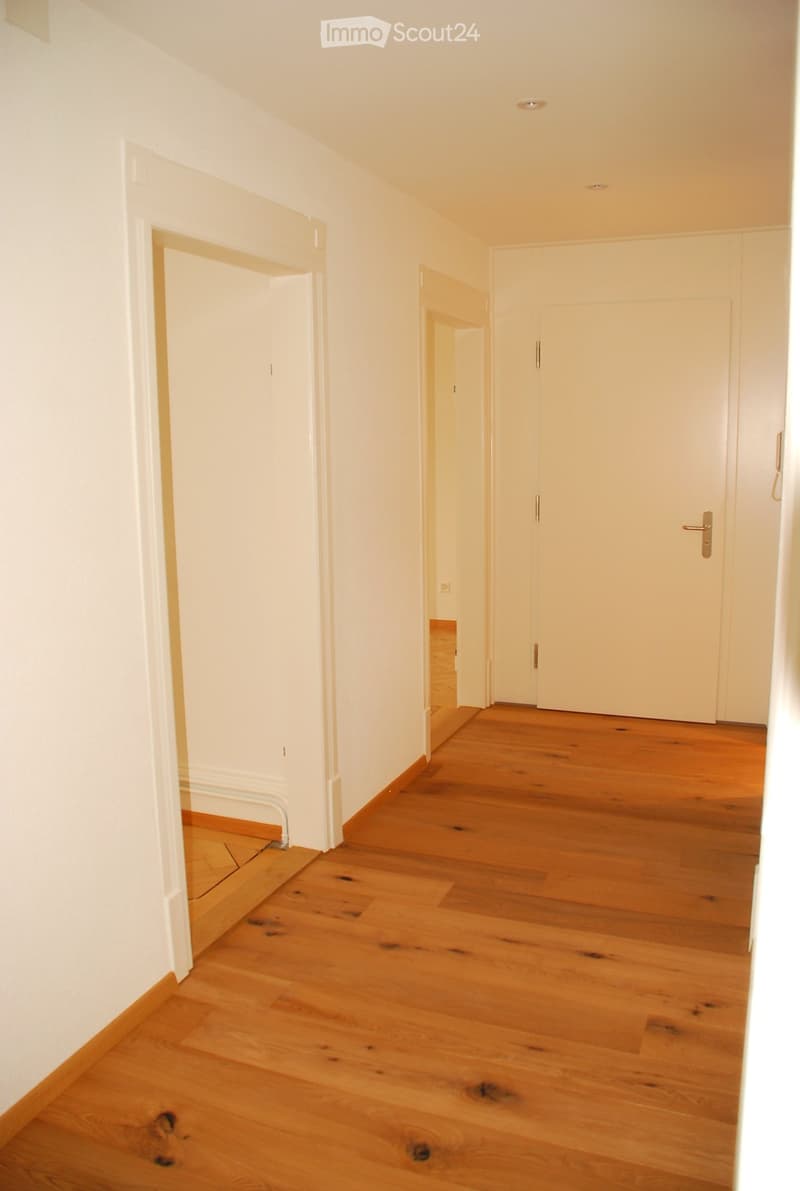 A clean, well-lit hallway with wooden flooring and white walls, featuring several doors and a visible light fixture on the ceiling.