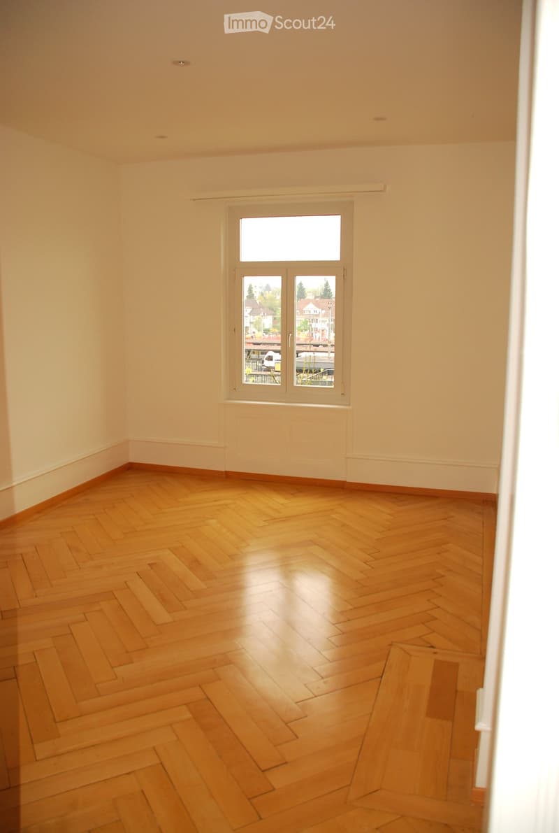 Empty room with wooden herringbone flooring, white walls, and a large window with a view of the outside.