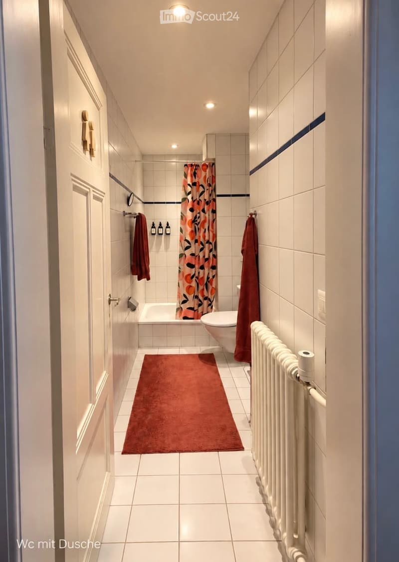 Modern bathroom with white tiled walls and floor, featuring a bathtub, toilet, radiator, towels, and a colorful shower curtain.