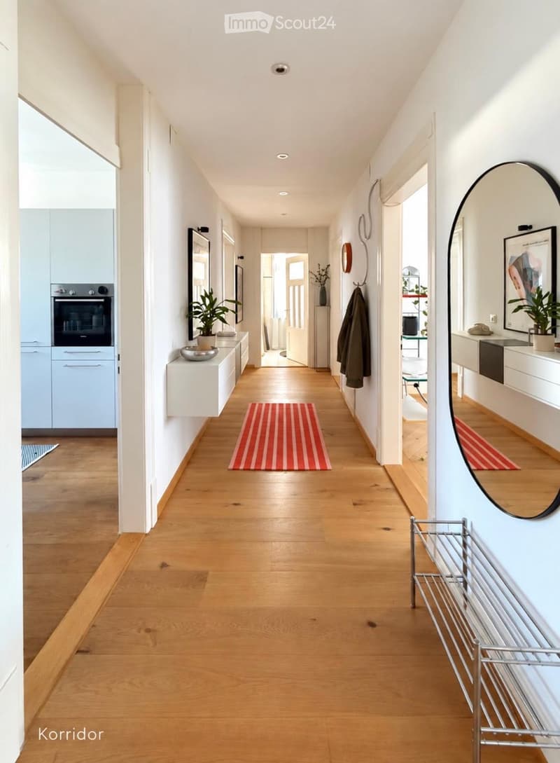 Bright hallway with wooden flooring, white walls, recessed lighting, and decorative elements including a round mirror, potted plants, and a coat rack.