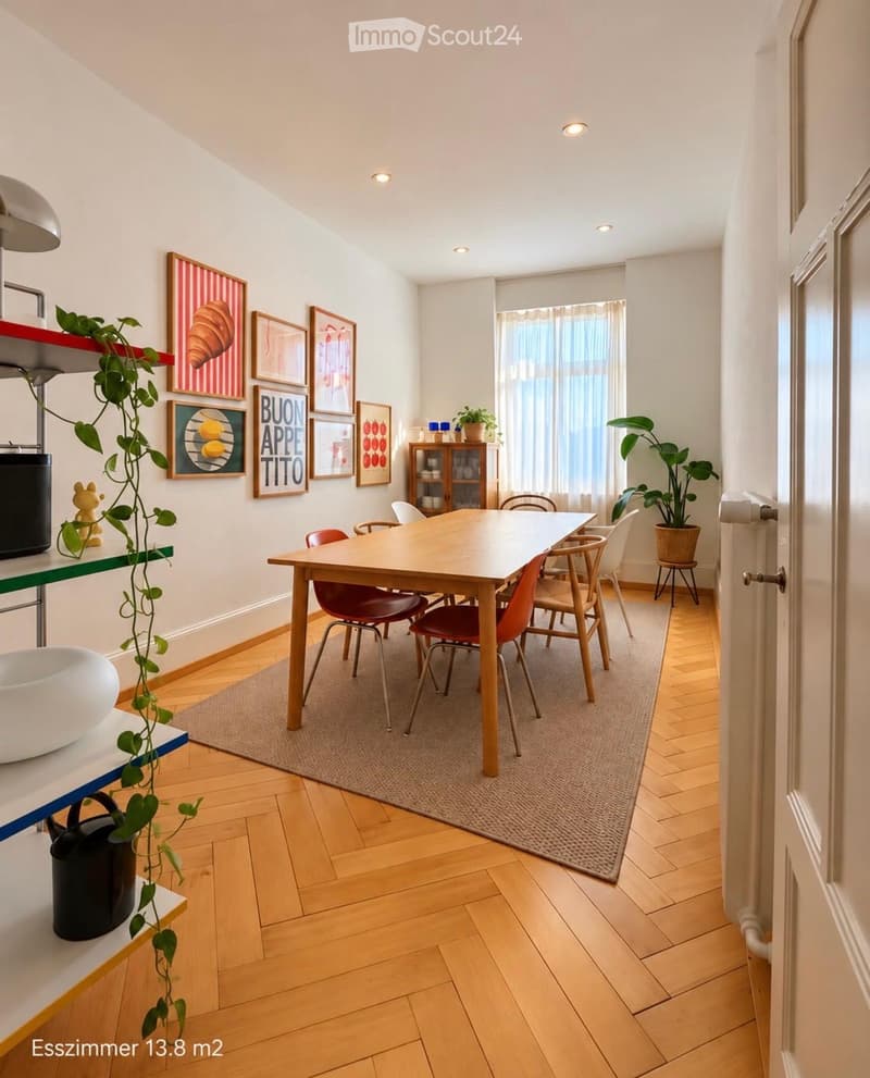 Bright kitchen with wooden flooring, a dining table surrounded by chairs, a small cabinet, potted plants, and framed artwork on the walls.