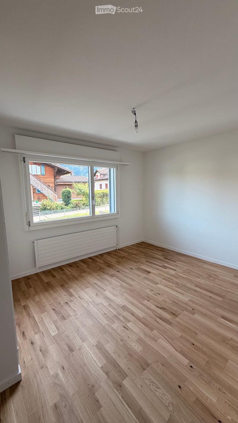 Empty room with light wooden flooring, white walls, large window with view of a house and garden, radiator, and a single ceiling light fixture.
