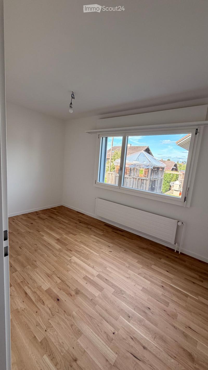 Empty room with light wooden flooring, white walls, large window with view of outdoor area, radiator, and single light fixture.