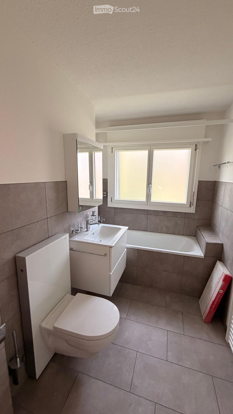 Modern bathroom with a toilet, sink, bathtub, mirror, and cabinet. Features tiled walls and a window with blinds.