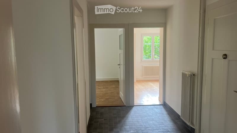 Empty hallway with tiled floor, wooden flooring in adjacent rooms, radiators, and natural light from a window in the adjacent room.