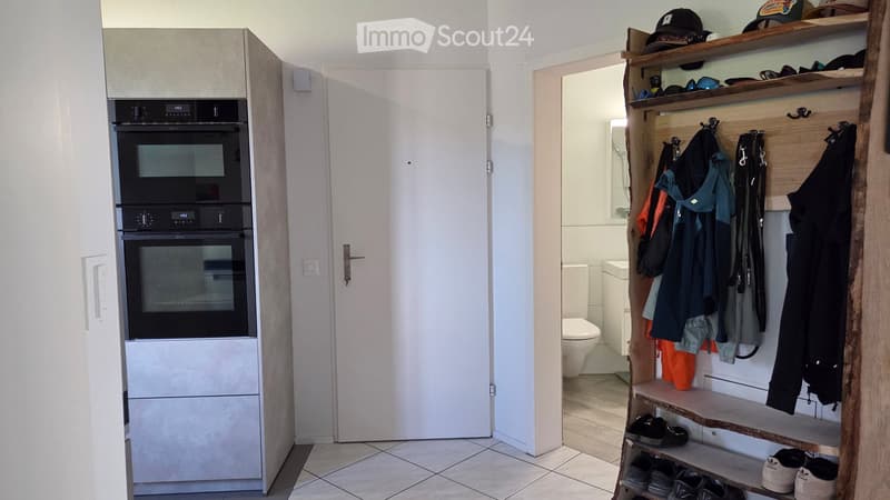 A modern, compact kitchen area with built-in ovens and a small bathroom visible through an open doorway. A wooden coat rack with shelves holds hats, coats, and shoes.