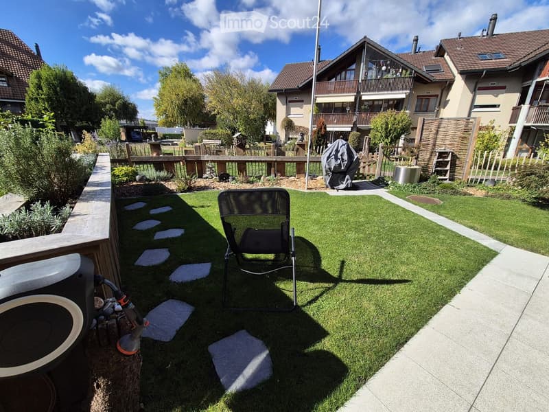 Small garden with artificial grass, a folding chair, and a hose reel. Wooden decking and stone stepping stones are visible.