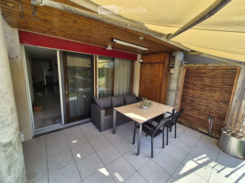 Balcony with tiled floor, wooden ceiling, sliding glass doors leading to interior, sofa, dining table with chairs, and potted plants.