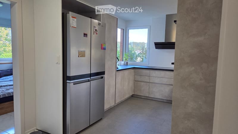 Modern kitchen with light-colored cabinets, stainless steel refrigerator, and a countertop with a small plant. The kitchen features a range hood and is connected to a bedroom area visible through an open doorway.