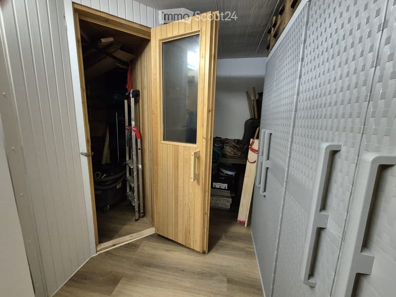 A narrow hallway with wooden flooring, a partially open wooden door leading to a storage area, and a grey storage unit on the right side.