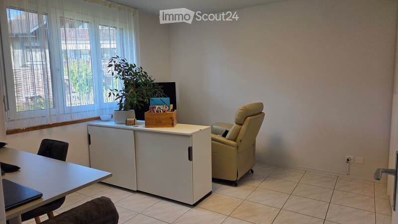 A furnished room with a tiled floor, a white table, a chair, a laptop, a small refrigerator, and a plant on the windowsill.