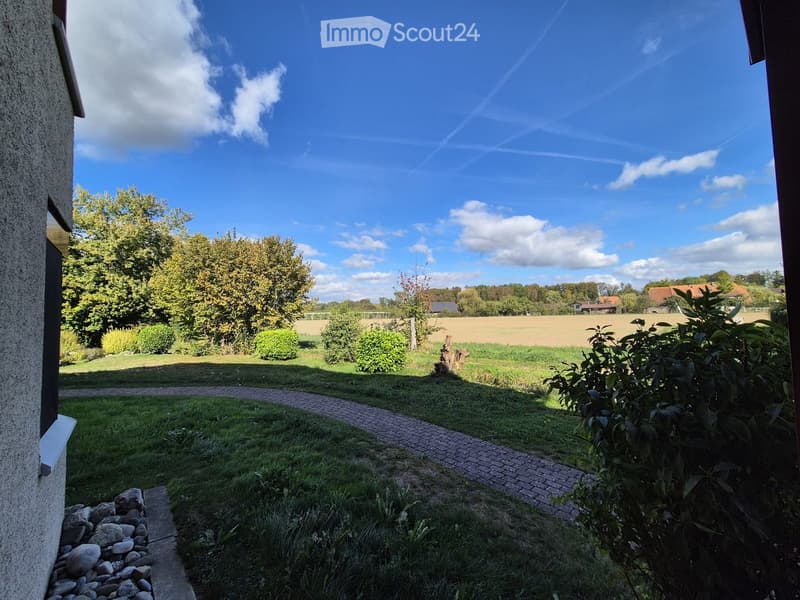Garden with a brick pathway, grassy area, trees, bushes, and a view of a field and distant houses.