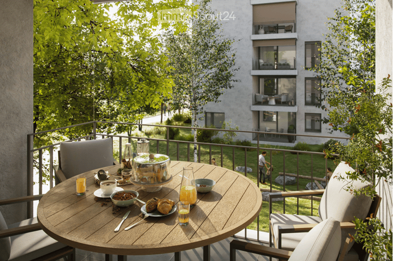 Balcony with a round wooden table set for breakfast, surrounded by chairs and a railing, overlooking a green park and modern apartment building.