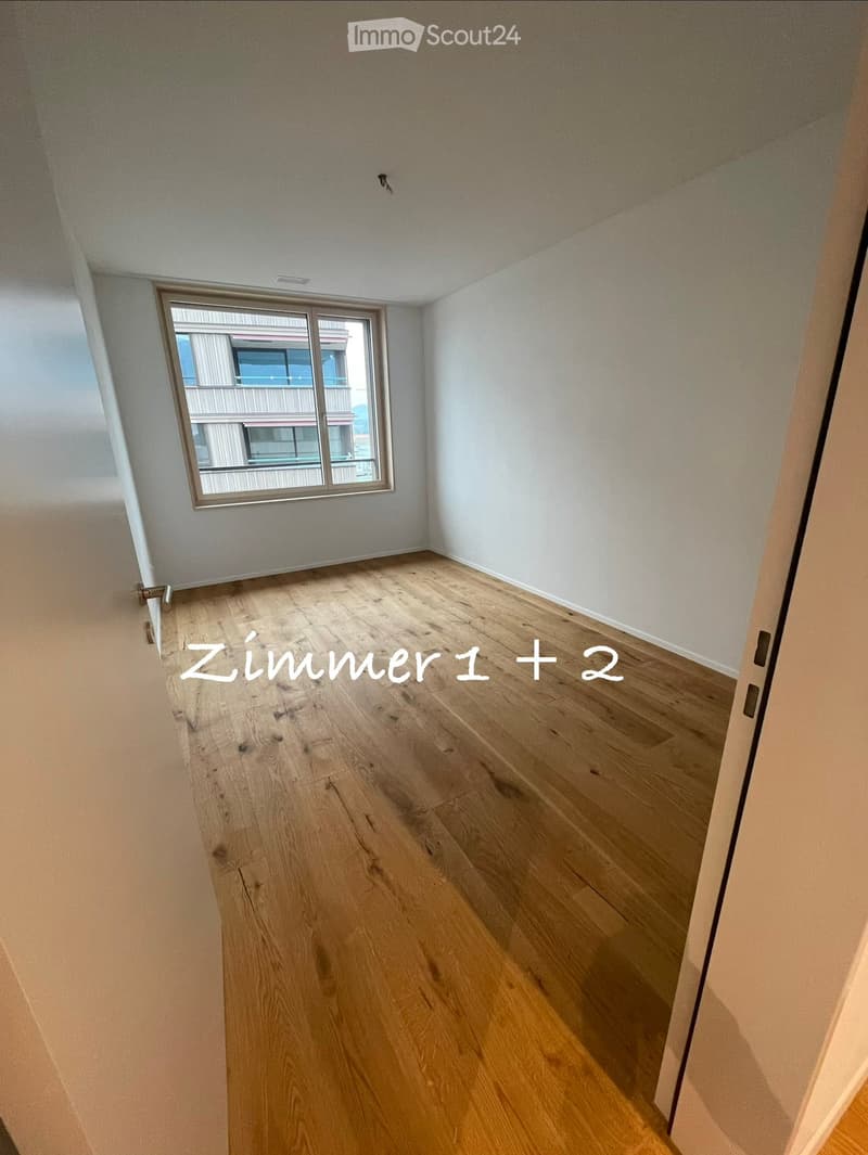 Empty room with wooden flooring, white walls, and a window with a view of another building.
