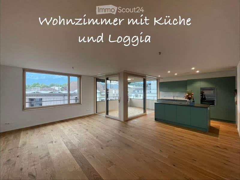 Open-plan living and kitchen area with wooden flooring, large windows, and a balcony access. Kitchen features green cabinets, island, sink, and decorative vase.
