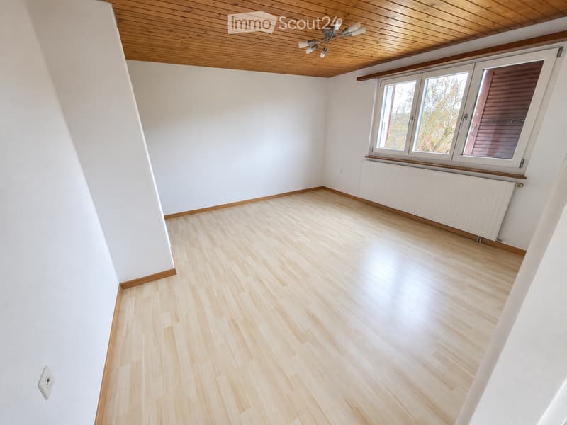 Empty room with light wooden flooring, white walls, and large windows with wooden frames. The ceiling is wooden with a central light fixture.