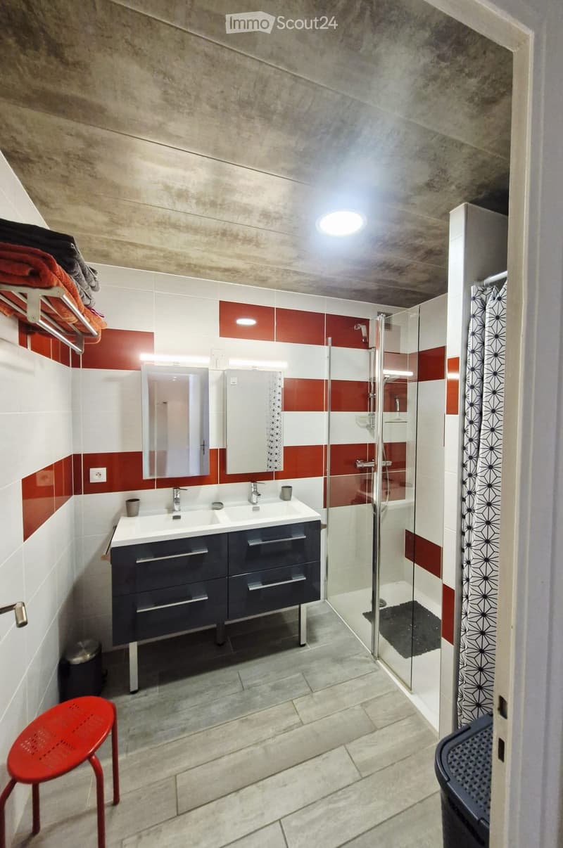 Modern bathroom with double sink, dark blue cabinet, red and white tiles, glass shower enclosure, and a red stool.