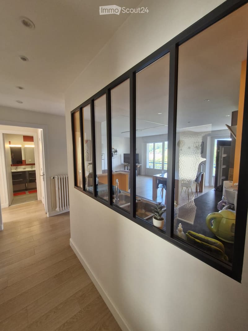 A modern hallway with wooden flooring, white walls, and large glass windows offering views into adjacent rooms, including a kitchen and living area.