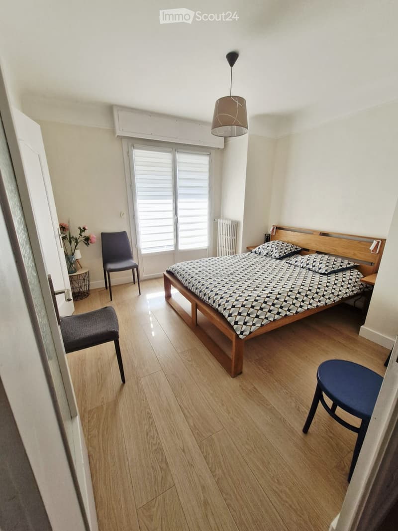 Cozy bedroom with wooden flooring, a double bed with patterned bedding, two chairs, a small table with a flower vase, and a hanging lamp.