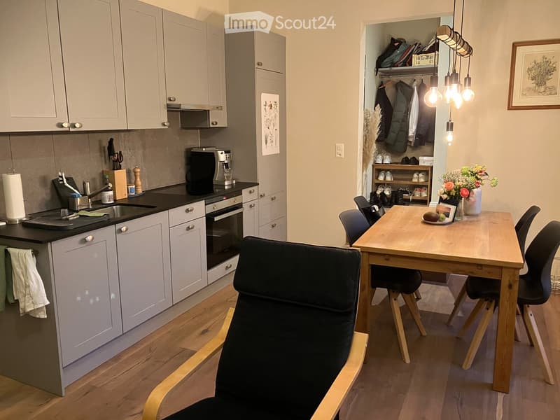 Modern kitchen with gray cabinets, black countertops, a large dining table, pendant lights, an oven, and a coffee machine.