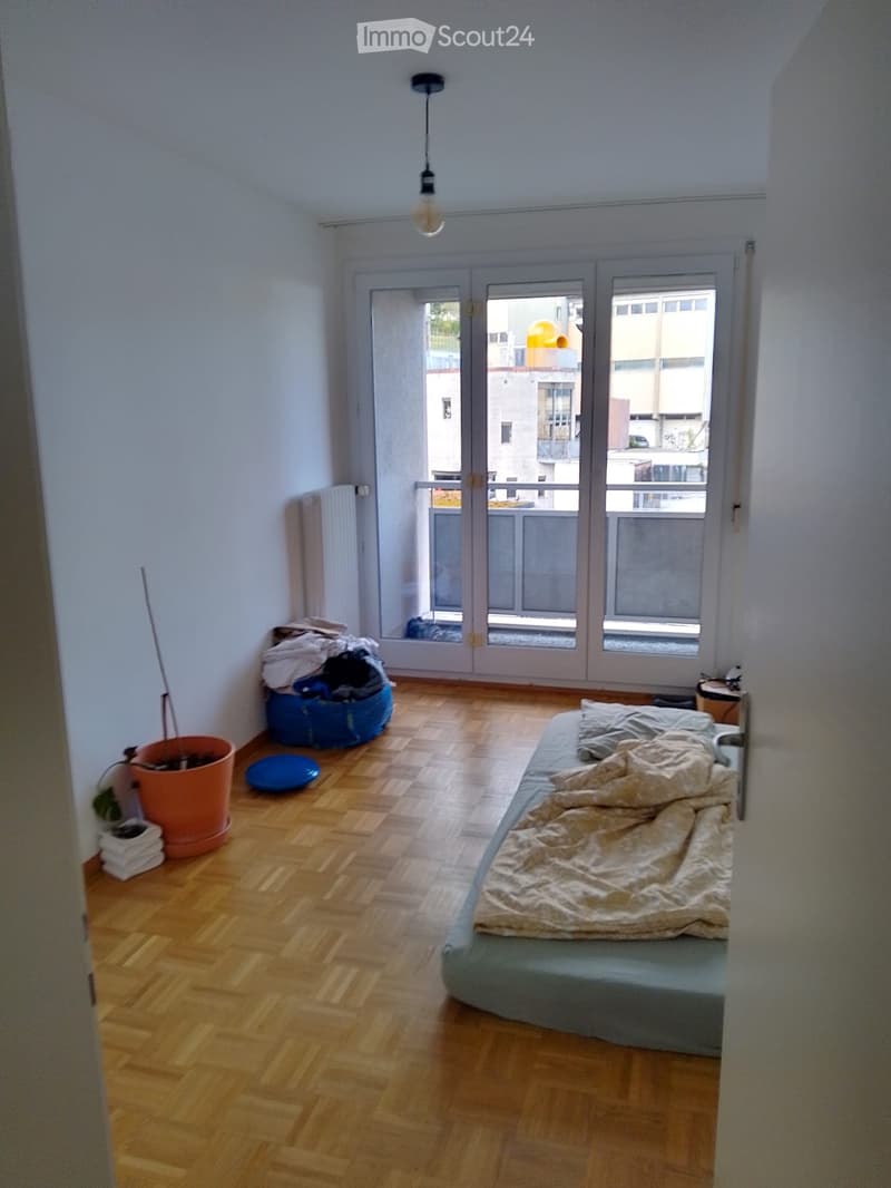 Bedroom with wooden floor, bed, radiator, potted plant, and a view through large windows.