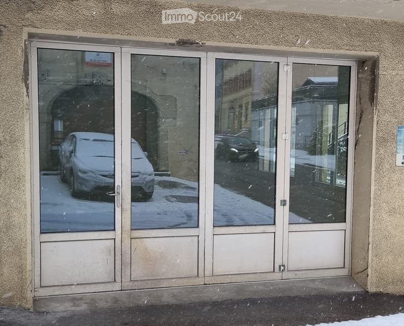 Entrance to a building with large glass doors, snow on the ground, and reflections of parked cars visible through the glass.