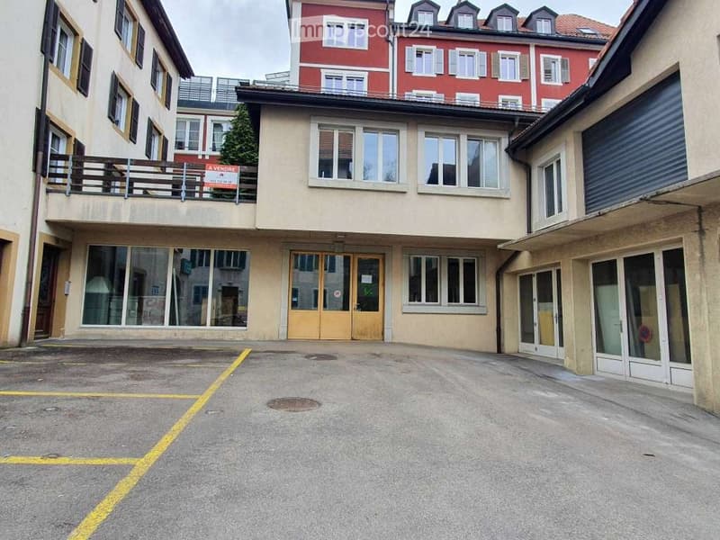 Multi-story building with a beige facade, large windows, and a balcony on the upper floor. The ground floor has glass storefronts and a main entrance with double doors.