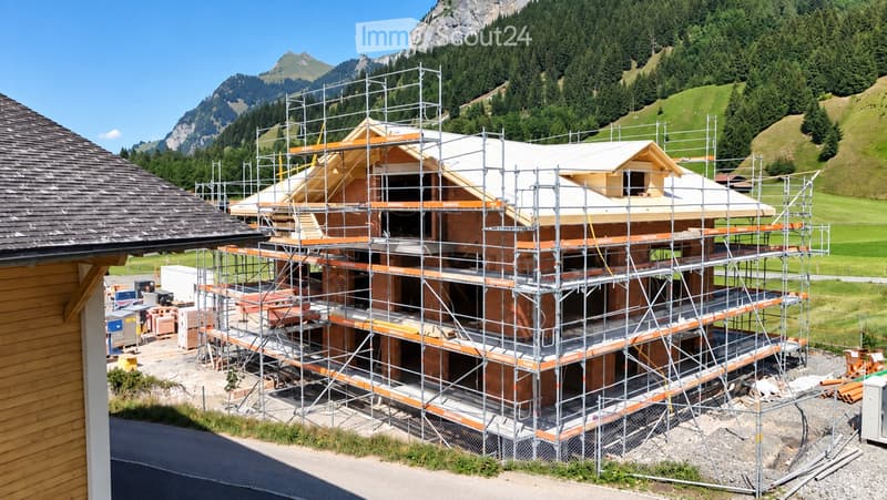 A multi-story house under construction, covered in scaffolding, with visible wooden beams and brickwork. The surrounding area features a grassy field and mountain backdrop.