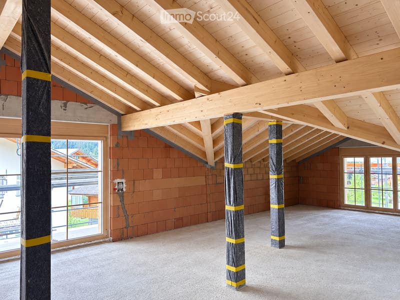 An empty attic space with exposed wooden beams and pillars, unfinished walls, and large windows offering a view of the surrounding area.