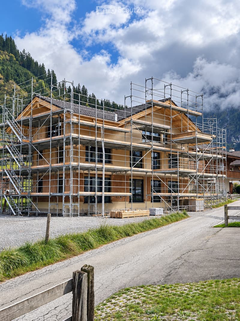A two-story house under construction, covered in scaffolding, with wooden framing and large windows, situated in a mountainous area with a gravel driveway and surrounding greenery.