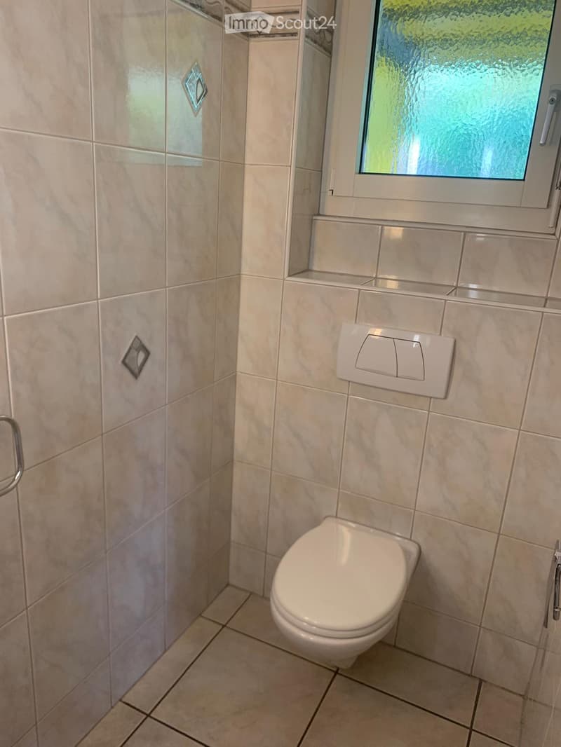 Modern bathroom with beige tiled walls and floor, a toilet, toilet paper holder, and a window with frosted glass.