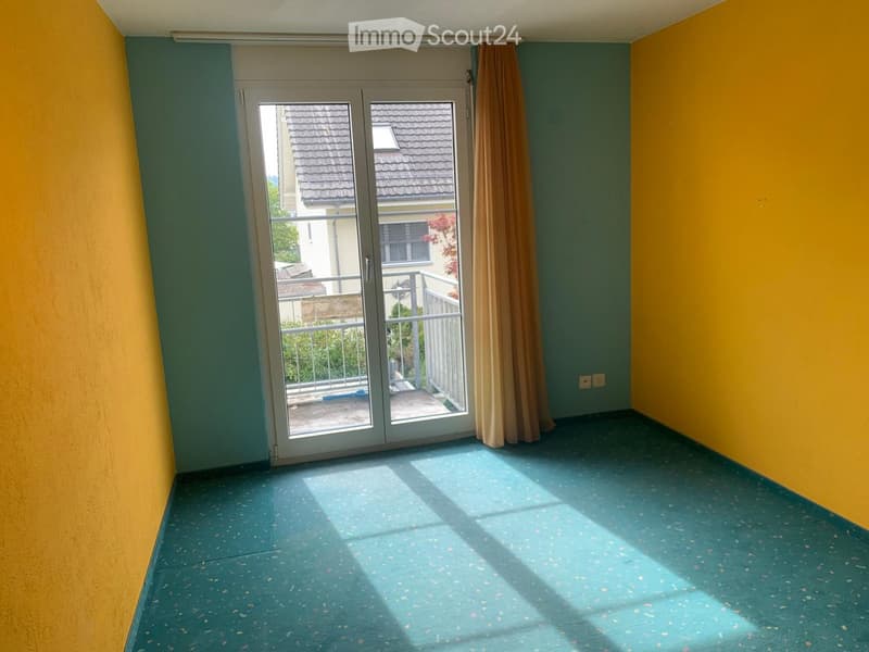 Empty room with blue speckled carpet, yellow and green walls, large sliding glass door leading to a balcony with a view of neighboring buildings.