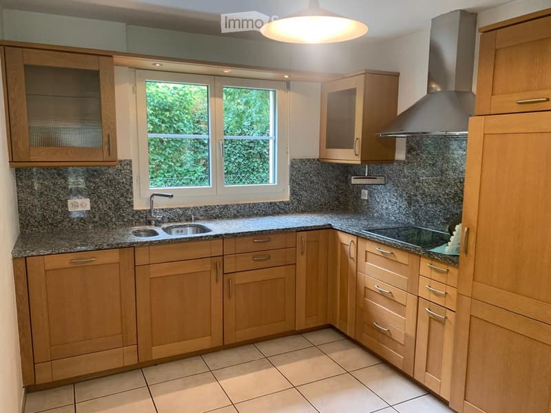 Modern kitchen with light wooden cabinets, granite countertops, a sink with a faucet, and an electric stove with a range hood. The room features tiled flooring and a large window with a view of greenery.