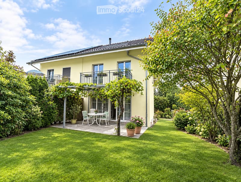 A well-maintained garden with a lawn, a patio area with a table and chairs, potted plants, and a house with a balcony and large windows.