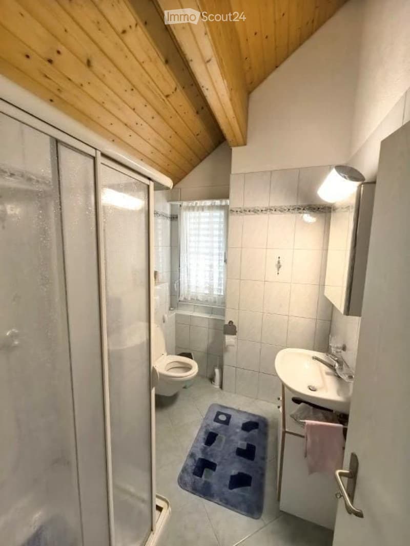Cozy bathroom with a toilet, sink, mirror, and a glass-enclosed shower. The room features a wooden slanted ceiling and a small blue rug on the floor.