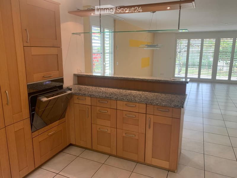 Modern kitchen with light wooden cabinets, granite countertop, built-in oven, and pendant lights. Large windows provide natural light.