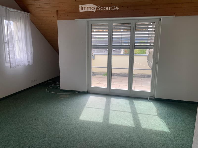 Empty room with green carpet, white walls, wooden slanted ceiling, large sliding glass doors leading to a balcony, and a window with white curtains.