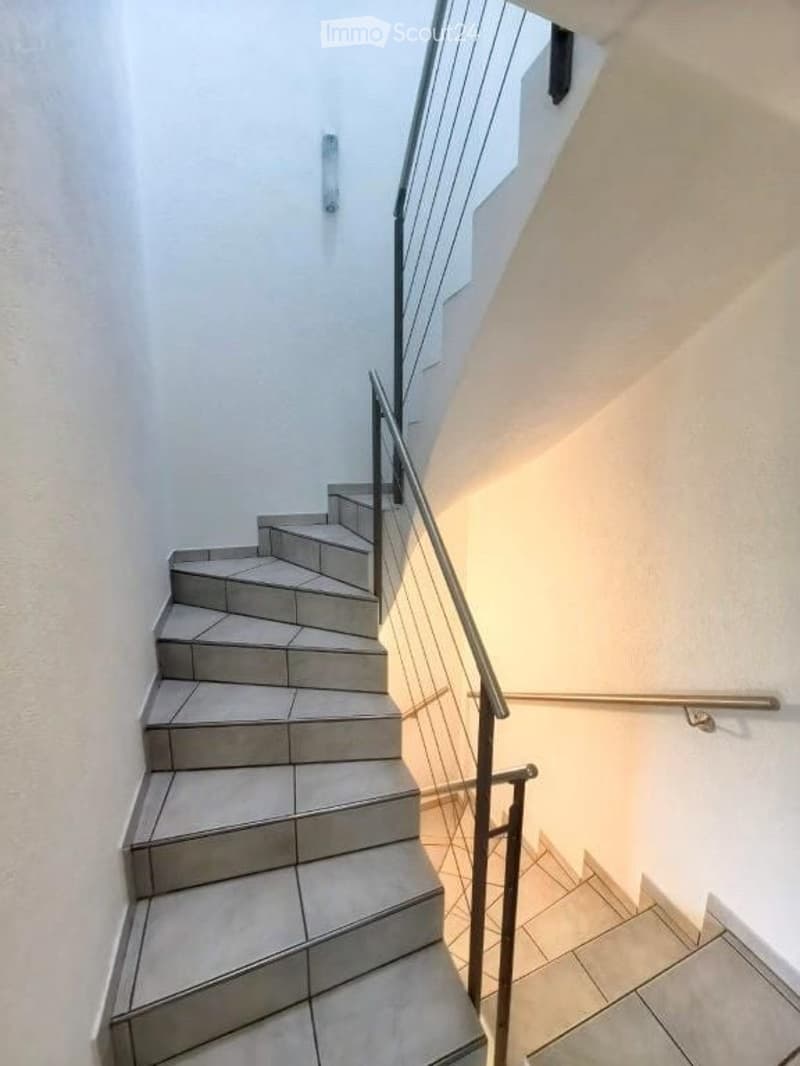 A modern staircase with gray tiled steps and a sleek metal railing, illuminated by natural light from a window at the top.