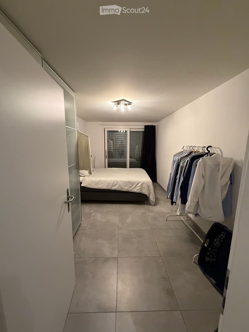 A modern bedroom with a bed, a wardrobe, and a clothing rack. The room features tiled flooring and large windows with blinds.