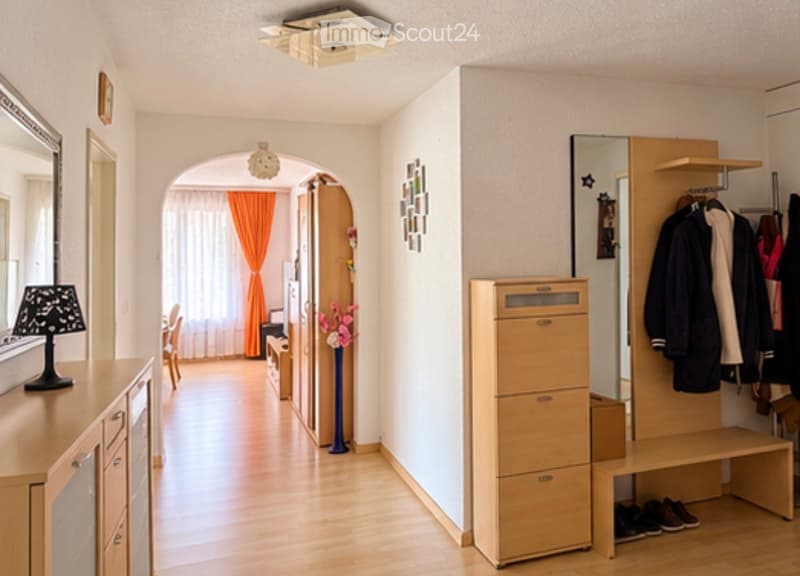 Bright hallway with wooden flooring, built-in storage unit, coat rack, and a view into an adjacent room with orange curtains.