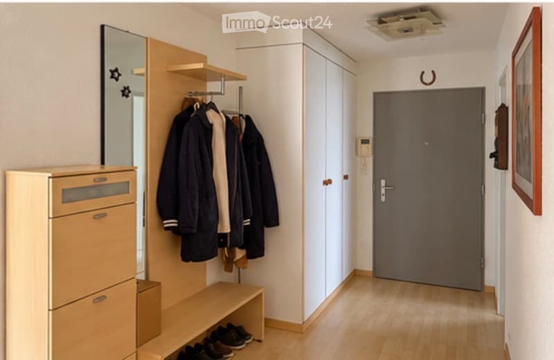 Modern hallway with light wooden flooring, built-in closet with mirrored doors, coat rack with jackets, small bench with shoes, and a grey door leading to another room.